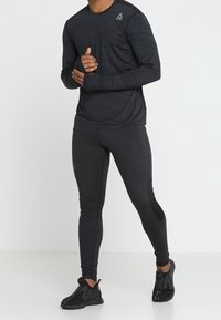 Mężczyzna w czarnej długiej koszulce sportowej, czarnych legginsach i czarnych trampkach, stojący na tle zwykłego białego tła.