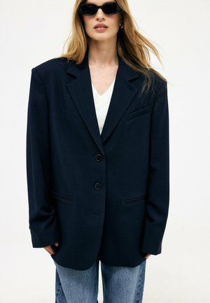 Donna con occhiali da sole neri, blazer scuro oversize, top bianco e jeans blu sta contro uno sfondo chiaro e semplice.