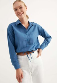 Camicia blu a maniche lunghe con bottoni, dotata di taschino sul petto, design cropped e dettagli a cravatta in vita, abbinata a pantaloni bianchi a vita alta.