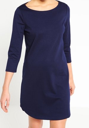 Woman wearing a simple, navy blue, knee-length dress with three-quarter sleeves and a round neckline.
