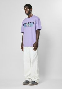Lilla bomull t-skjorte med grafisk design på forsiden, kombinert med hvite vide jeans og grå sneakers med klare såler.