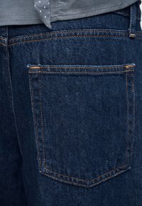 Poche arrière d'un jean en denim bleu foncé avec des coutures orange, porté avec une chemise grise montrant un coin noué à motifs de pois blancs.