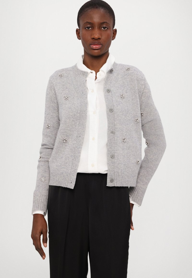 Graue Strickjacke mit Perlenverzierungen, gerippten Bündchen und Saum, runder Ausschnitt und Knopfverschluss, über einem weißen Hemd getragen.
