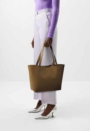 Brown leather tote bag with two slender handles, smooth texture, and a wide rectangular shape, held by a person in light purple attire.