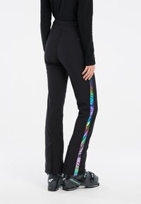 Zwarte skibroek met een multicolore iriserende zijkant, een slim fit en ritsen bij de enkels. Gecombineerd met zwarte skischoenen.