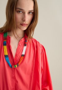 Camicia corallo brillante con dettagli in bottone, caratterizzata da una collana di perline multicolori con motivi vivaci, appesa al collo.