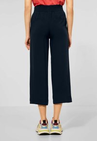 Marinblå vida byxor med smockad midja, med två bakfickor och slätt tyg. Modellen har kombinerat dem med färgglada sportskor.