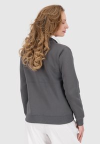 Donna con lunghi capelli ricci biondi, indossa una felpa grigia e pantaloni bianchi, rivolta leggermente di lato contro uno sfondo neutro.