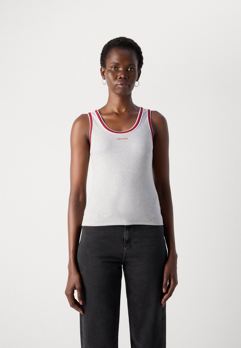 Frau, die frontal steht und ein graues Tanktop mit rotem Saum und schwarze Jeans trägt, vor einem einfachen weißen Hintergrund.