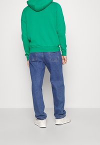 Persona che indossa un felpa verde, jeans blu e sneakers bianche in piedi di spalle contro uno sfondo bianco semplice.