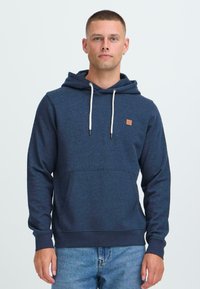 Giovane con capelli corti che indossa un hoodie blu navy con lacci bianchi e jeans azzurri, in piedi davanti a uno sfondo bianco semplice.