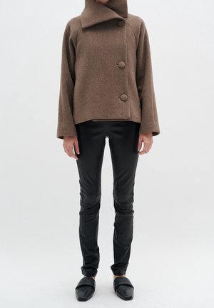 Persoon draagt een bruine asymmetrische wollen blousejas met knopen, zwarte aansluitende leren broek en zwarte instapschoenen op een lichte achtergrond.