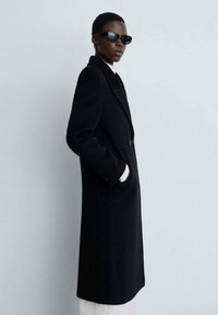 Zwarte wollen jas met een reverskraag, sluiting met één knoop, zijzakken en witte gebreide manchetten. Model draagt zwarte zonnebril.