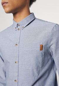 Camicia blu chiaro con bottoni in tessuto intrecciato, dotata di colletto, bottoni marroni e taschino sul petto con etichetta in pelle.