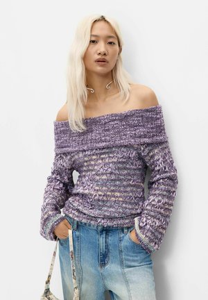 Jeune femme aux longs cheveux blonds portant un pull texturé violet tombant sur les épaules et un jean bleu clair, les mains dans les poches.
