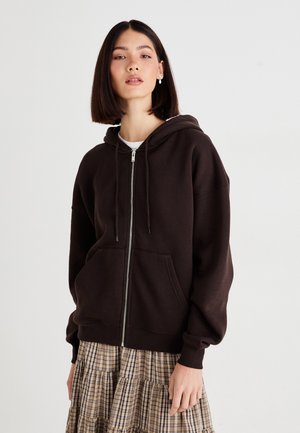 Brown zip-up hoodie with a drawstring hood, loose fit, and front pocket, complemented by a plaid skirt. Cotton material with soft texture.