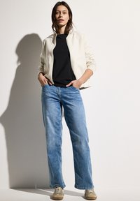 Ljust beige zip-up jacka, svart t-shirt och blå bredbeniga jeans. Sneakers i en beige nyans, släta tygstrukturer, avslappnad design.