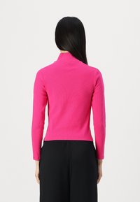 Růžový vypasovaný svetřík s vysokým límcem, dlouhými rukávy, strukturovanou tkaninou a zkráceným střihem, kombinovaný s černými, volně střiženými kalhotami.