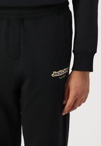 Fekete melegítőnadrág puha anyagból, oldalsó zsebekkel és sárga "Jack & Jones" logóval a bal lábon. 