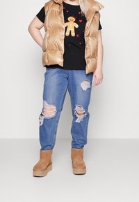 Osoba ubrana w beżową kamizelkę puchową, czarny t-shirt z nadrukiem piernika, przetarte niebieskie dżinsy i beżowe buty na platformie, stojąca na tle jednolitym.