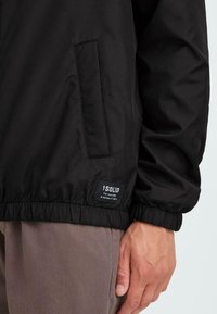 Black lightweight jacket, featuring a zip pocket and elastic cuffs. Includes a label at the hem with brand name and tagline.