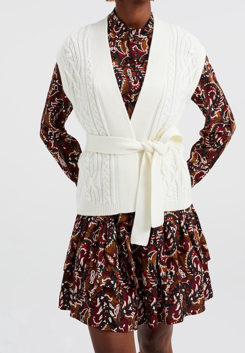 Gilet blanc en tricot avec ceinture à nouer, superposé à une robe brune à motifs floraux avec volants. Les matériaux semblent doux et texturés.