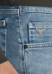 Vaqueros de mezclilla azul claro con un bolsillo trasero prominente que lleva un logo plateado con alas y un botón negro, combinados con un suéter oscuro de punto acanalado.