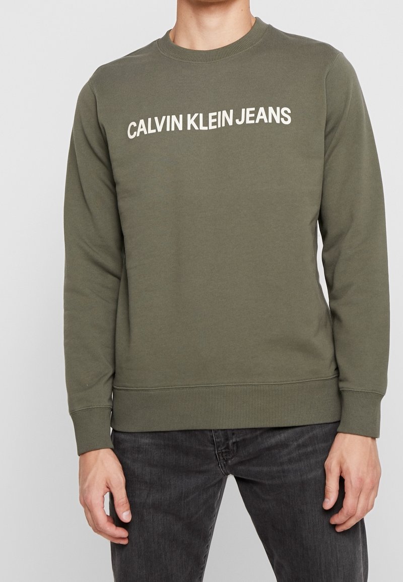 Felpa verde oliva in cotone, con collo a giro, polsini a coste e la scritta "CALVIN KLEIN JEANS" stampata in bianco sul petto.