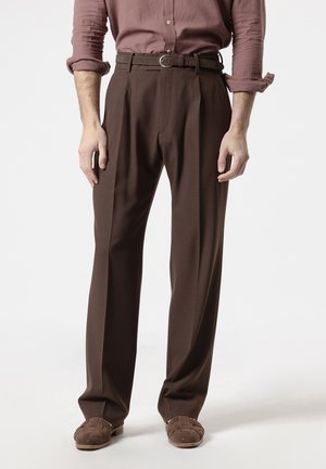 Hombre con pantalones marrones plisados, cinturón a juego, zapatos marrones de ante tejidos y camisa de manga larga remangada de pie sobre fondo blanco.