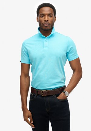 Hellblaues Polohemd mit umgeschlagenem Kragen, kurzen Ärmeln und einer Knopfleiste. Trägt dazu eine dunkle Jeans und einen braunen Gürtel. Silberne Armbanduhr sichtbar.