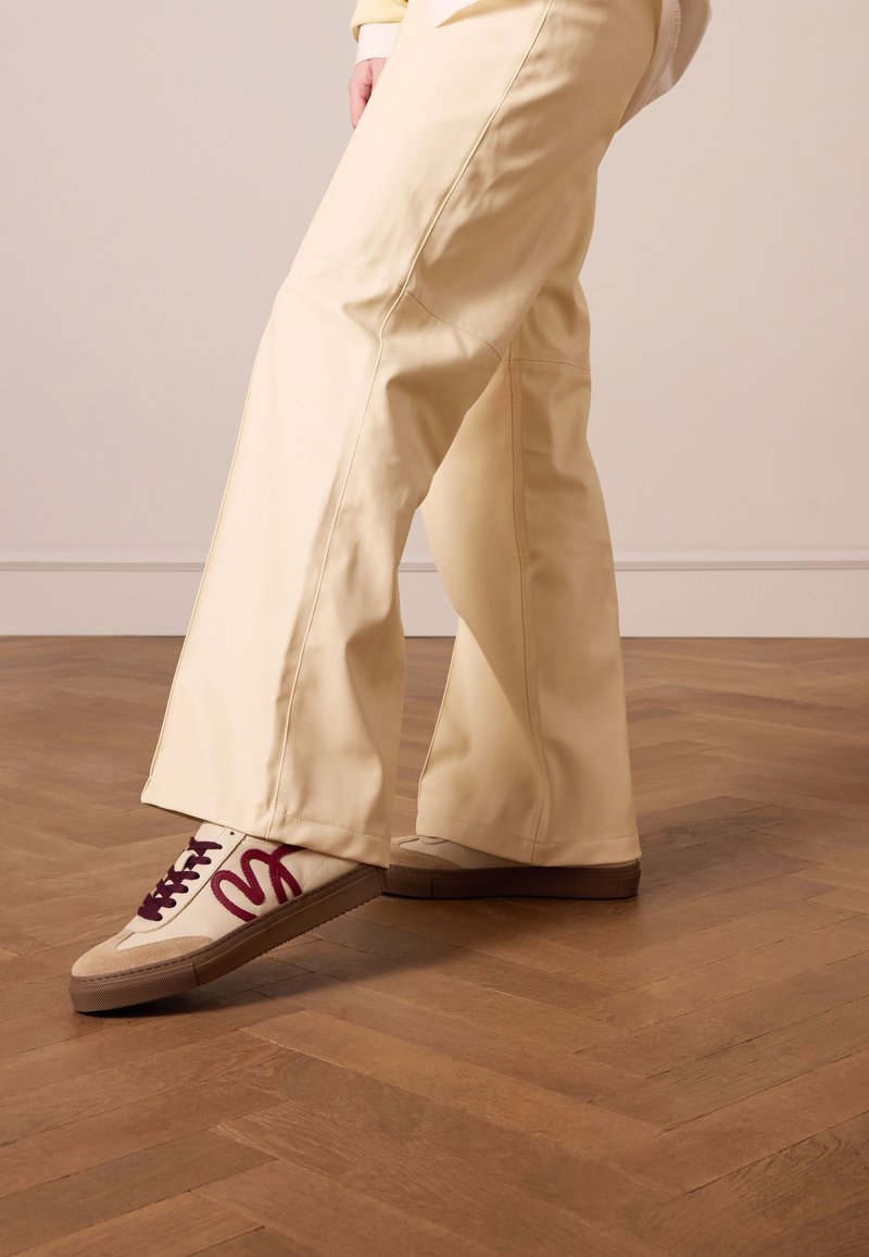 Witte sneakers met kastanjekleurige accenten en donkere veters, gecombineerd met crèmekleurige brede broek van gladde stof, op een houten vloer.