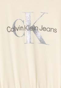 Krémová tkanina Calvin Klein Jeans s grafickým nápisem 'CK' a textem 'Calvin Klein Jeans' v šedé barvě, s jemnou texturou a stříbrným glitrem.