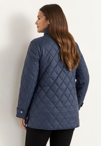 Marineblaue gesteppte Jacke mit hohem Kragen, Knopfdetail an den Manschetten und strukturiertem Rautenmuster. Trägt sie über schwarzen Hosen.