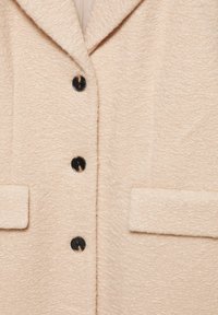 Beżowy płaszcz o fakturze z wyłogowym kołnierzem, z czterema dużymi czarnymi guzikami i dwiema przednimi kieszeniami. Miękka, puszysta tkanina.
