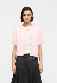 Γυναίκα που φοράει ανοιχτό ροζ κοντομάνικο ζακέτα πάνω από λευκό πουκάμισο με κόκκινη καρδιά, σε συνδυασμό με ψηλόμεση μαύρη πλισέ φούστα.