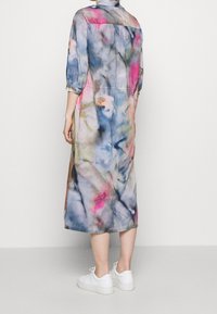 Kobieta w luźnej, midi sukience tie-dye z rękawami do 3/4 i białych trampkach, stojąca na tle jednolitego, jasnego tła.