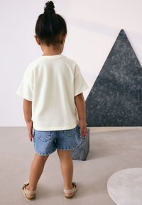 Cream short-sleeve t-shirt with a relaxed fit, paired with blue denim shorts. Background includes geometric shapes in muted colors.