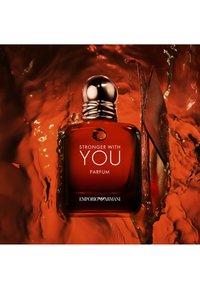 Bruine glazen parfumfles met een ronde metalen dop, voorzien van witte tekst "STRONGER WITH YOU PARFUM" en accentuerend met oranje vloeistof en een lederen detail.