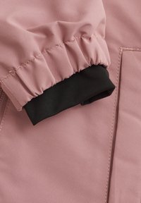 Rosa jacka i tyg med samlade muddar. Svart ribbstickad knit på den inre mudden. Slät textur med synlig sömnad och subtil lyster.