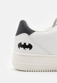 Weißer Sneaker mit schwarzem Fersenakzent, seitlich mit einem schwarzen Batman-Logo, strukturierter Gummisohle und gesteppten Details.