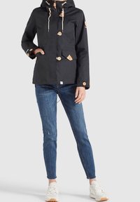 Veste noire imperméable avec capuche, boutons en bois et deux poches frontales, portée avec un jean bleu et des baskets blanches.