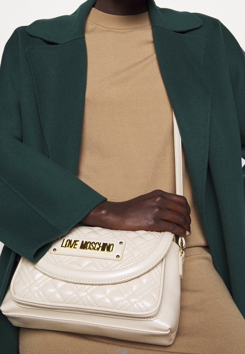 Beige laukku, jossa on tikattu pinta ja kultainen "LOVE MOSCHINO" -plakaatti, irrotettavalla olkahihnalla, tyylitelty tummanvihreän takin ja vaaleanruskean paidan kanssa.