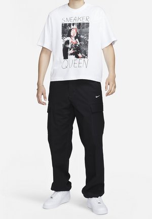 Osoba ubrana w białą koszulkę z grafiką "Sneaker Queen", czarne spodnie typu cargo z logo Nike oraz białe trampki, stojąca na białym tle.