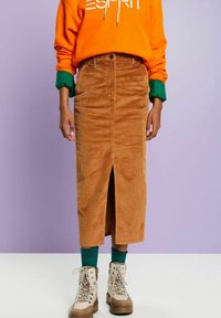 Brauner Cord-Midirock mit einem Schlitz vorne, getragen mit einem orangefarbenen Hoodie und grünen Akzenten. Verfügt über eine taillierte Taille und gerippte Textur.