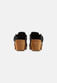 Sabots en suede noir avec un talon en bois, présentant un design découpé et des accents en forme de nœud de chaque côté, vus de l'arrière.
