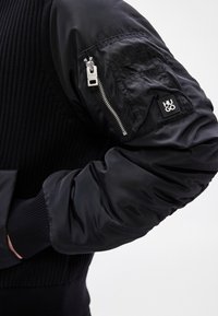 Casaco bomber preto com corpo em malha canelada, mangas fofas e lisas, bolso com fecho de correr no braço esquerdo e um emblema do logotipo.