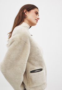 Krēms krāsas mākslīgās ādas jaka ar augstu apkakli un platām piedurknēm. Iezīmējas ar melniem akcentiem uz kabatām, nodrošinot maigu, pūkainu tekstūru.