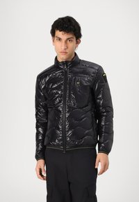 Giacca puffer nera con colletto alto, design trapuntato, texture lucida, zip frontale e tasca sul petto. Presenta un accento verde sull'orlo.