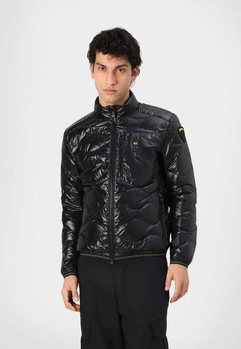 Giacca puffer nera con colletto alto, design trapuntato, texture lucida, zip frontale e tasca sul petto. Presenta un accento verde sull'orlo.