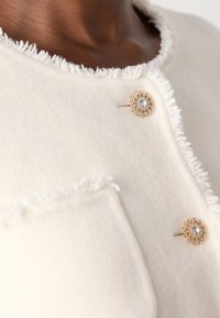 White wool jacket featuring a fringed neckline, two gold flower-shaped buttons with rhinestones, and a single front pocket.